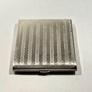 May include: A silver-colored, rectangular cigarette case with a textured surface. The case features vertical striped patterns and a decorative border. The clasp is visible on the bottom edge. The case is likely made of metal.