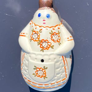 Puede incluir: Una figurita de cerámica blanca de una mujer con un moño marrón y un vestido blanco con diseños florales naranja y marrón. La figurita tiene ojos azules y una boca roja.