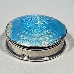 May include: A small, round silver box with a bright blue, patterned enamel lid. The lid features a detailed geometric design. The box has a silver base and sides, with hallmarks visible. A decorative item.