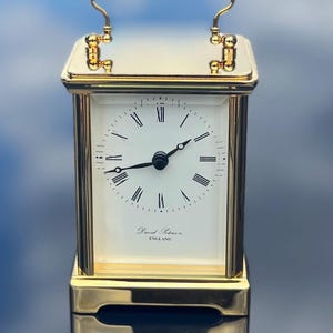 May include: A gold-tone carriage clock with a white face, black Roman numerals, and black hands. The clock features a top handle and the text David Peterson ENGLAND printed on the dial. The polished metal frame has a reflective, smooth finish.