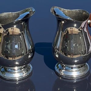 Puede incluir: Dos jarras plateadas con asas de cuero marrón. Las jarras tienen un cuerpo bulboso y un labio acampanado. Las asas están unidas a la parte superior de la jarra y se curvan hacia abajo para encontrarse con el cuerpo. Las jarras están sobre una superficie azul oscuro.