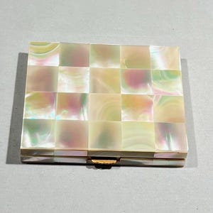 May include: A white and iridescent mother of pearl compact with a checkered pattern. The compact has a gold metal clasp.