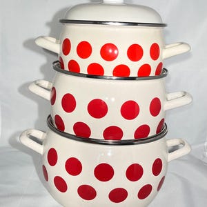 May include: A set of three white enamel pots with red polka dots. The pots are stacked on top of each other, with the largest pot at the bottom and the smallest pot at the top. The pots have silver rims and handles.