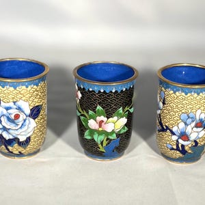 Peut inclure: Trois petits gobelets cylindriques décoratifs avec des bords bleus et des motifs floraux. Les gobelets sont en métal et ont une finition en émail cloisonné. Les motifs présentent des fleurs dans des tons de bleu, blanc et jaune sur un fond noir ou crème.