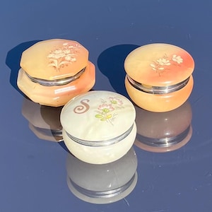 Puede incluir: Tres pequeñas cajas redondas decorativas de alabastro. Las cajas están pintadas con diseños florales y tienen tapas plateadas. Una caja tiene la letra "S" pintada.