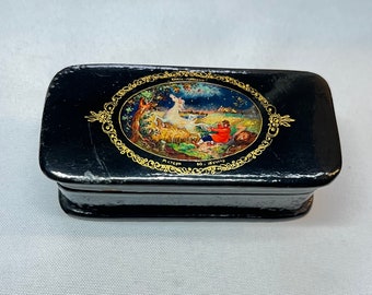 May include: A black lacquered wooden box with a gold-trimmed oval on the lid. The oval features a painted scene of a woman and a man in a forest setting. The text "Meister  Ю. Жуков" is written in gold lettering below the oval.