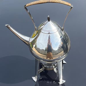 May include: A silver teapot with a woven handle sits on a stand with three legs. The teapot is polished and reflects the surrounding environment.