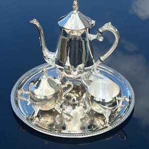 May include: A polished silver tea set consisting of a teapot with a lid, a lidded sugar bowl, and an open creamer, all featuring ornate handles and feet. The three pieces are arranged on a round silver serving tray with a beaded edge detail.