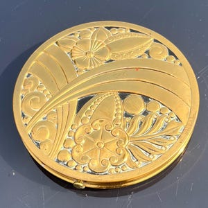 Op de afbeelding: Een gouden compact spiegel met een Art Deco-ontwerp met een bloemenpatroon. De spiegel heeft een zwarte achtergrond en een gouden rand.