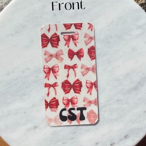 Puede incluir: Una etiqueta de equipaje rectangular con un patr&oacute;n de lazos rojos sobre un fondo blanco. La etiqueta tiene las letras "CST" en negro. La etiqueta est&aacute; sobre una superficie de m&aacute;rmol con una base de madera.