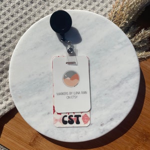 Puede incluir: Un portatarjetas blanco con una parte superior circular negra y una correa de pl&aacute;stico transparente. La tarjeta presenta el texto "MARKERS BY LUNA RAIN ON ETSY" y "CST" en una fuente decorativa, con un patr&oacute;n floral.