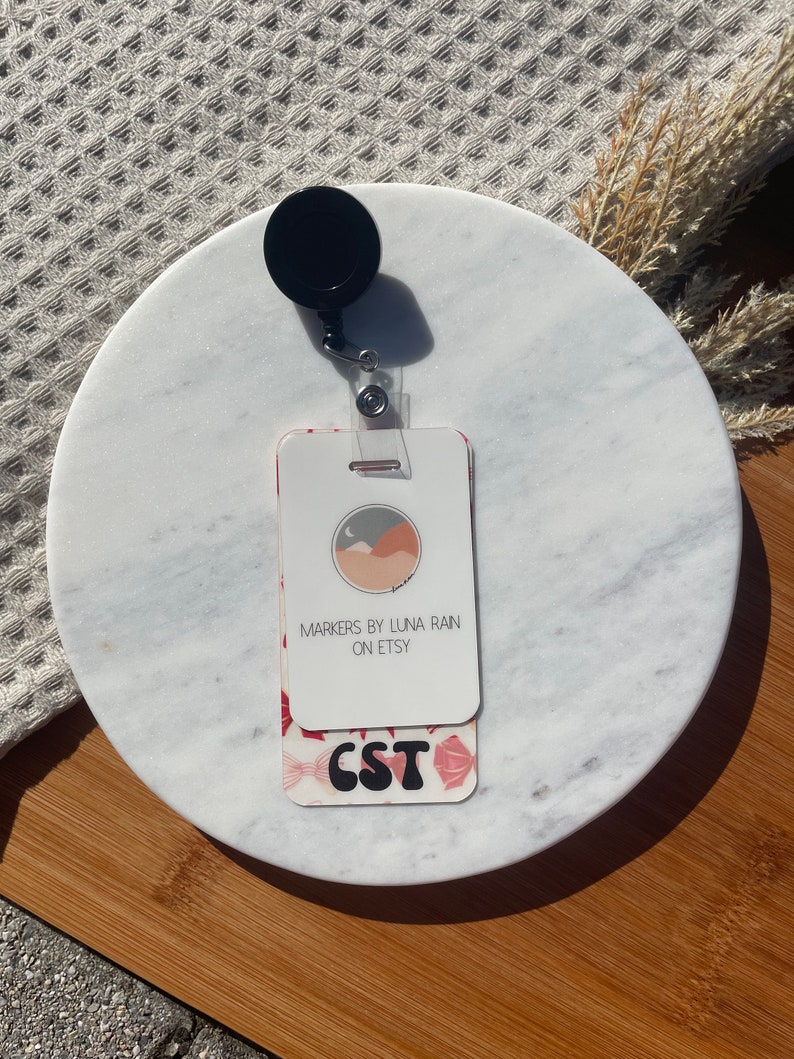 Puede incluir: Un portatarjetas blanco con una parte superior circular negra y una correa de pl&aacute;stico transparente. La tarjeta presenta el texto "MARKERS BY LUNA RAIN ON ETSY" y "CST" en una fuente decorativa, con un patr&oacute;n floral.