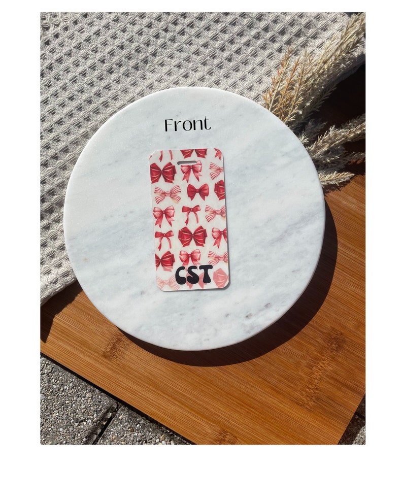 Puede incluir: Una etiqueta de equipaje rectangular con un patr&oacute;n de lazos rojos sobre un fondo blanco. La etiqueta tiene las letras "CST" en negro. La etiqueta est&aacute; sobre una superficie de m&aacute;rmol con una base de madera.