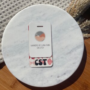 Puede incluir: Un portatarjetas blanco y rojo con el texto "CST" y "MARKERS BY LUNA RAIN ON ETSY". El portatarjetas tiene un dise&ntilde;o floral y un peque&ntilde;o gr&aacute;fico de monta&ntilde;a y luna. El portatarjetas est&aacute; sobre una superficie de m&aacute;rmol blanco.