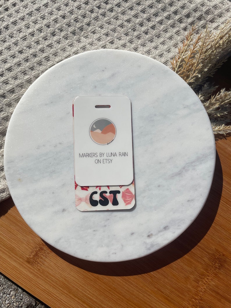 Puede incluir: Un portatarjetas blanco y rojo con el texto "CST" y "MARKERS BY LUNA RAIN ON ETSY". El portatarjetas tiene un dise&ntilde;o floral y un peque&ntilde;o gr&aacute;fico de monta&ntilde;a y luna. El portatarjetas est&aacute; sobre una superficie de m&aacute;rmol blanco.