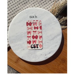 Puede incluir: Una etiqueta rectangular con un patr&oacute;n de lazos rojos sobre un fondo crema. La etiqueta tiene un agujero en la parte superior y las letras "CST" en una fuente retro negra en la parte inferior. La palabra "Back" est&aacute; impresa en la superficie de m&aacute;rmol.