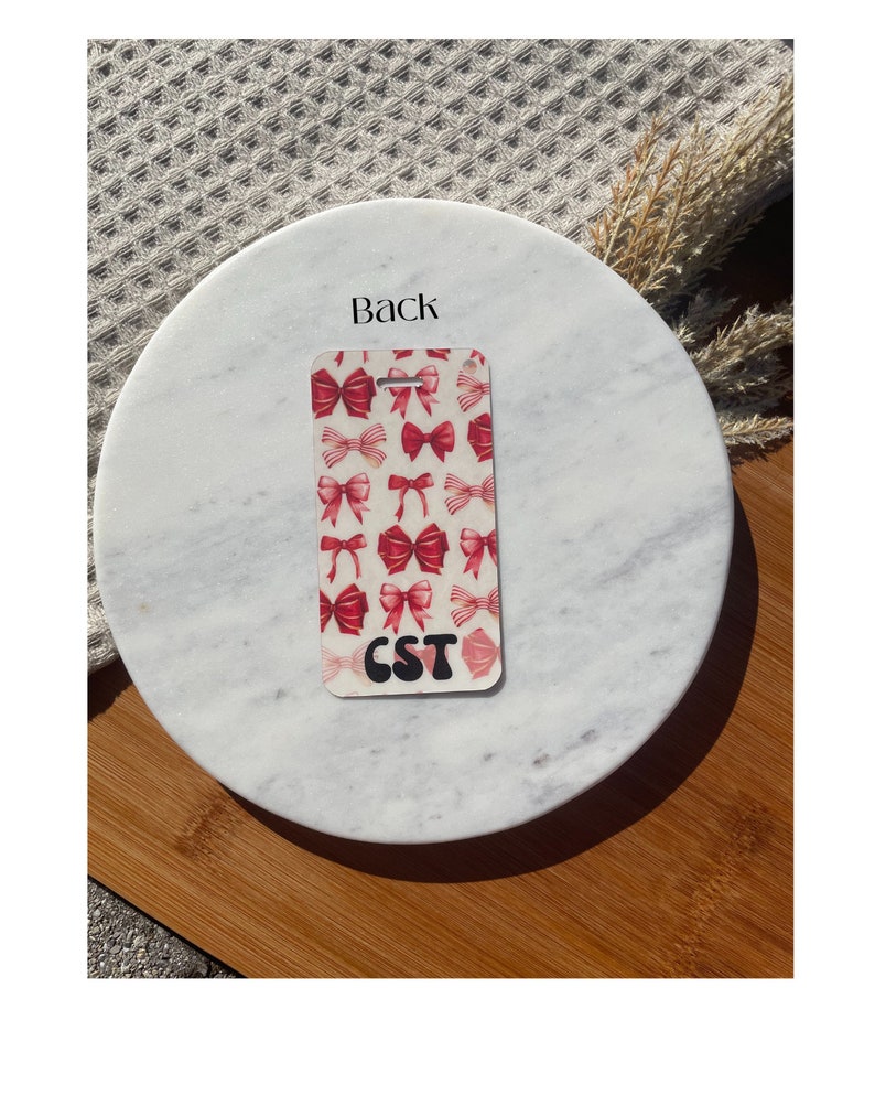 Puede incluir: Una etiqueta rectangular con un patr&oacute;n de lazos rojos sobre un fondo crema. La etiqueta tiene un agujero en la parte superior y las letras "CST" en una fuente retro negra en la parte inferior. La palabra "Back" est&aacute; impresa en la superficie de m&aacute;rmol.