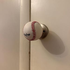 Op de afbeelding: Een witte honkbal met rode stiksels en het zwarte Rawlings-logo op de oppervlakte, gemonteerd als een functionele deurknop. De honkbal is bevestigd aan een ronde basisplaat van geborsteld zilverkleurig metaal die aan de deur vastzit.