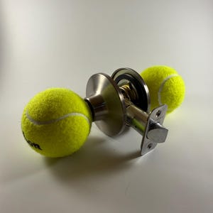 May include: A metal door knob assembly featuring two bright neon yellow tennis balls attached to the ends of the handle. The mechanism includes a circular brushed silver plate, a central latch bolt, and internal brass-colored components.