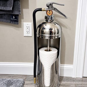 May include: A polished chrome toilet paper holder designed to resemble a fire extinguisher. It features a vertical cylindrical body with a cutout for a roll of white toilet paper, a black rubber hose, and a silver metal handle mechanism.