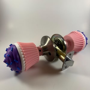 May include: A door knob set featuring two cupcake-shaped handles. Each handle has a pink pleated base, a purple top, and small blue sprinkles. The central mechanism is made of brushed silver metal with a standard latch and mounting plate.