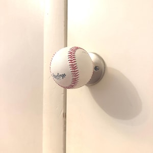 May include: A white baseball with red stitching and the black Rawlings logo printed on the surface, mounted onto a circular brushed metal base attached to a white door. The baseball functions as a door knob, featuring visible red laces.