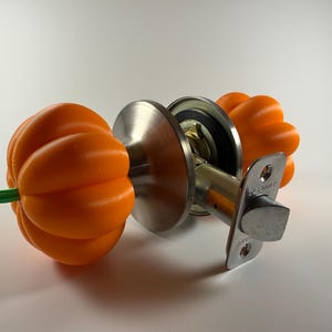 May include: A door knob set featuring two bright orange, pumpkin-shaped handles with small green stems. The central mechanism is made of brushed silver-tone metal, including a circular plate and a rectangular latch plate with the text Defiant.