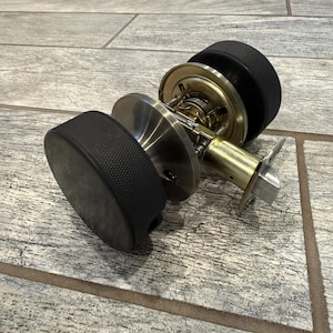Könnte beinhalten: Ein Türknauf-Set bestehend aus zwei schwarzen, texturierten Eishockey-Pucks, die an einer metallischen Basis in gebürsteter Nickeloptik befestigt sind. Der Mechanismus umfasst einen messingfarbenen Zylinder, eine Federfalle und ein Metallblech.