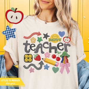 May include: Cream-colored t-shirt with a colorful "Happy Teacher" design. The design includes a pencil, stars, apples, cherries, and a rainbow. The word "teacher" is in a black and white checkered pattern.
