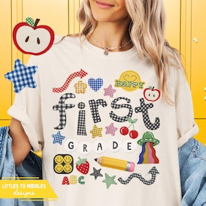 May include: Cream-colored t-shirt with the words "First Grade" in a playful font, decorated with colorful patches of stars, hearts, apples, and a pencil. The shirt has a whimsical design, perfect for back-to-school.