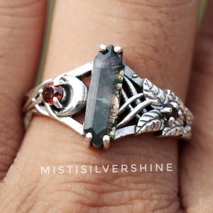 May include: A silver ring featuring a rectangular, dark green gemstone. The ring has a detailed floral and moon design with a small red gemstone accent. The text "MISTISILVERSHINE" is visible on the ring.