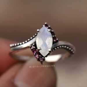 May include: A silver ring featuring a marquise-cut, pearly white gemstone, accented with small, dark red gemstones. The ring's band has a dotted design. The ring is held in a hand, with the brand name "MISTISILVERSHINE" visible.