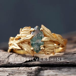 May include: A gold ring with a central, teardrop-shaped stone with green and white inclusions. The ring's band and setting feature a leaf and floral design. The text "MISTISILVERSHINE" is visible at the bottom of the image.