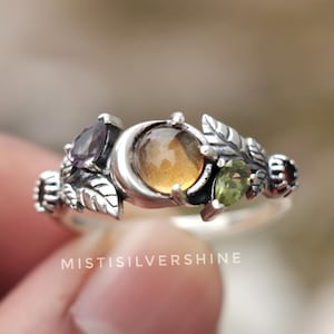 Könnte beinhalten: Ein silberner Ring mit einem Mond-Design und drei Edelsteinen. Der Ring hat einen bernsteinfarbenen Mittelstein, flankiert von einem lila und einem grünen Stein, mit Blatt- und Rankendetails. Der Text "MISTISILVERSHINE" ist sichtbar.