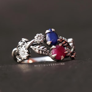 May include: A silver ring with a floral design featuring three gemstones: a blue sapphire, a red ruby, and a smaller white stone. The ring is set on a dark background.