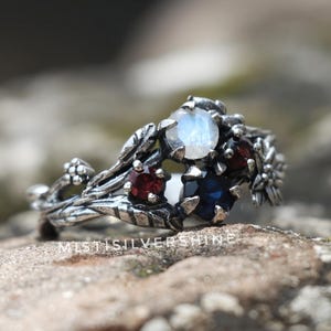 Könnte beinhalten: Silberring mit floralem Design, mit einem großen, runden, irisierenden Mondstein und kleineren roten und blauen Edelsteinen. Der Ring hat ein detailliertes, von der Natur inspiriertes Design mit Blättern und Blumen.