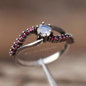 May include: A silver ring with a central, round, light blue gemstone, surrounded by smaller, red gemstones. The ring has a split band design. The text "MISTISILVERSHINE" is visible on the ring.