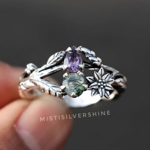 May include: A silver ring with a delicate floral design featuring two gemstones. The ring has a green and black gemstone and a purple gemstone.