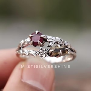 May include: A silver ring with a dark red gemstone at the center. The ring features a floral and leaf design, with the text "MISTISILVERSHINE" visible. The ring is held by a person's hand.