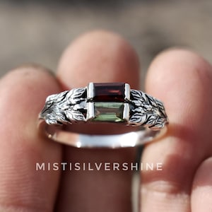 May include: A silver ring with a dark red and light green rectangular gemstone setting. The ring has a leaf design on the band and the text "MISTISILVERSHINE".