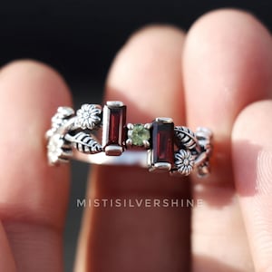 以下が含まれることがあります： 赤い宝石と緑の宝石がついた花柄の銀の指輪。指輪には小さな花と葉が飾られており、宝石は房状に配置されています。