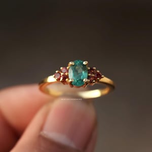 May include: A gold ring featuring an oval, teal-colored gemstone as the centerpiece, flanked by clusters of smaller, red gemstones. The ring has a delicate band and a classic design, suitable for various occasions.