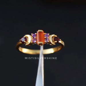May include: Gold ring featuring a rectangular orange gemstone and smaller purple and red gemstones. The ring is displayed against a white background.