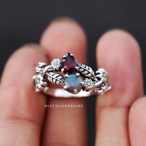 Könnte beinhalten: Ein silberner Ring mit floralem Design, der mit zwei Edelsteinen besetzt ist: einem roten tropfenförmigen Stein und einem runden, schillernden Stein. Der Ring sitzt an einem Finger.