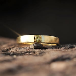 Pode incluir: Um anel de ouro com uma pedra preciosa quadrada verde claro no centro. O anel é exibido em uma superfície marrom áspera, com um fundo escuro. O anel tem uma banda retangular simples.
