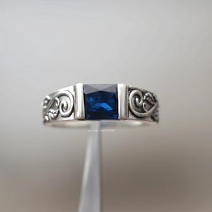 May include: A silver ring with a rectangular, deep blue gemstone. The band has decorative scrollwork on either side of the stone. The ring is shown against a soft, neutral background, highlighting its elegant design.