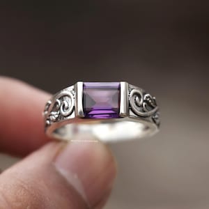 May include: A silver ring with an amethyst gemstone. The rectangular purple stone is set between two silver bars. The band features an ornate, swirling design. The ring is held up against a blurred background.