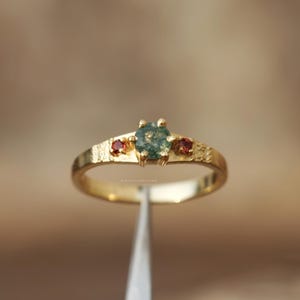 May include: A gold ring with a textured band features a central round, moss-green gemstone flanked by two smaller red gemstones. The ring's design is simple yet elegant, showcasing the natural beauty of the stones.