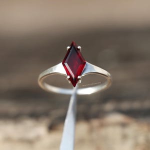 May include: A silver ring featuring a single kite-shaped, deep red gemstone held in a four-prong setting. The band is smooth and polished, tapering slightly as it meets the central stone. The text MISTISILVERSHINE is visible near the base of the setting.
