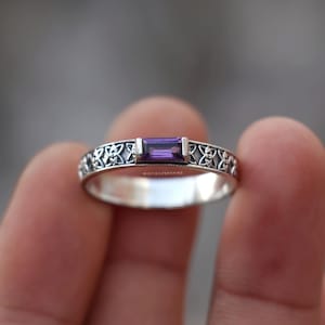 May include: A silver ring featuring a rectangular baguette-cut purple amethyst gemstone set in the center. The band is decorated with an intricate repeating knotwork pattern and includes the text MISTISILVERSHINE engraved on the inner surface.
